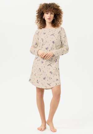 Vrouw met krullend haar die een lange mouw beige jurk met vogels-, maan- en sterrenpatronen draagt, staat op blote voeten op een witte achtergrond.