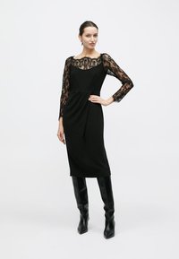 Robe noire avec manches longues en dentelle, corsage ajusté et jupe drapée. Portée avec de grandes bottes noires. Détail élégant d'encolure en dentelle visible.