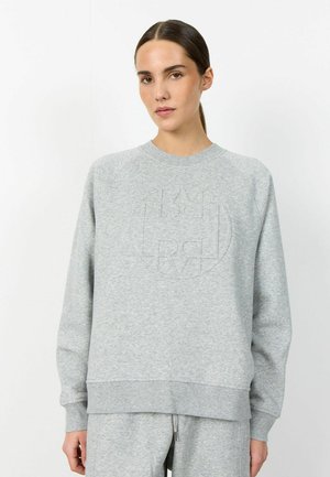 Frau mit braunem nach hinten gebundenem Haar, die einen hellgrauen Pullover und passende Jogginghose trägt und vor einem weißen Hintergrund steht.