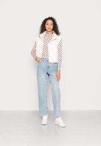 Colete acolchoado branco por cima de uma blusa de manga longa rosa com padrões de corações vermelhos. Jeans ligeiramente azuis de perna reta e ténis brancos. Roupa simples e casual.