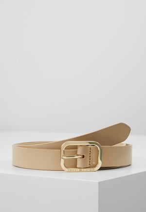 Beżowy skórzany pasek zwinięty na białej powierzchni z prostokątną złotą klamrą z wygrawerowanym napisem "Tommy Hilfiger".