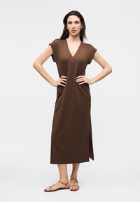 Frau steht mit den Händen in der Hüfte, trägt ein ärmelloses braunes Midikleid mit seitlichem Schlitz und flachen Sandalen vor einfarbigem Hintergrund.