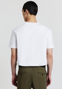 Uomo visto di spalle che indossa una maglietta bianca a maniche corte, semplice, infilata in un paio di pantaloni verde oliva con tasche posteriori con bottoni.