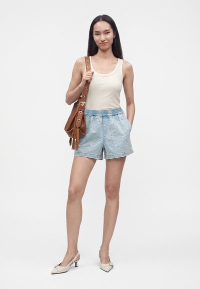 Jeune femme portant un débardeur beige, un short bleu clair à imprimé floral, des talons bas blancs et un sac à bandoulière marron avec des glands.