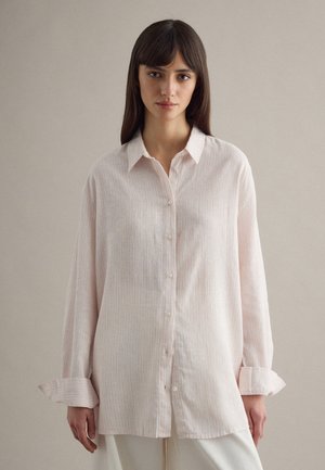 Vrouw met recht, donker haar draagt een losse, lichtroze overhemd met knopen en witte broek, staande tegen een effen beige achtergrond.