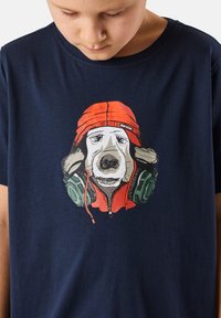 Laivansinis puuvillapaita, jossa on grafiikka koirasta, joka käyttää kuulokkeita ja punaista hupparia, yksityiskohtaisella sarjakuvamaisella suunnittelulla.