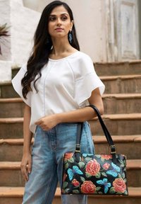 Zwarte handtas met levendige bloemen- en vlinderpatronen, zilverkleurige hardware en dubbele hengsels. Model draagt een witte top en hoge taille jeans.