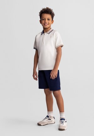 Garçon aux cheveux bouclés portant un polo blanc, un short bleu marine, des chaussettes blanches et des baskets blanches, debout et souriant sur un fond gris.