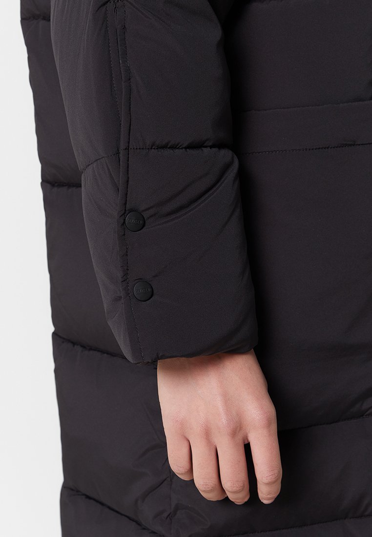 Schwarze Pufferjacke mit einem Ärmel, der eine glatte Textur aufweist, zwei schwarzen Knopfverschlüssen und horizontalen Steppnähten. Hand an der Manschette positioniert.