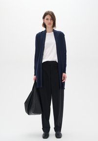 Marineblå lang cardigan laget av mykt stoff, sammen med en hvit skjorte og svarte løse bukser, bærende en stor svart læder tote bag.