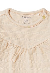 Lichtbeige babyshirt van textuurstof, met ronde hals en geplooide details onder de schouders. Bevat een kleine gouden knoop als accent.