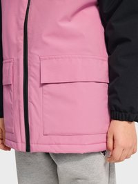 Kind trägt eine pinke Jacke mit schwarzen Ärmeln, Frontreißverschluss, großen Klapptaschen und grauer Hose, steht mit locker neben dem Körper hängender Hand.