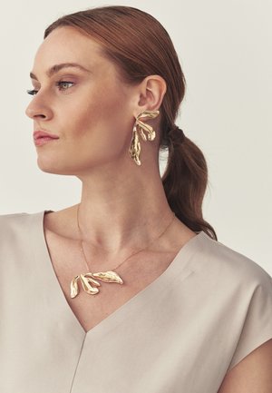 Woman with pulled-back hair wears large, gold, abstract leaf-shaped earrings and matching necklace, dressed in light beige top, facing left.