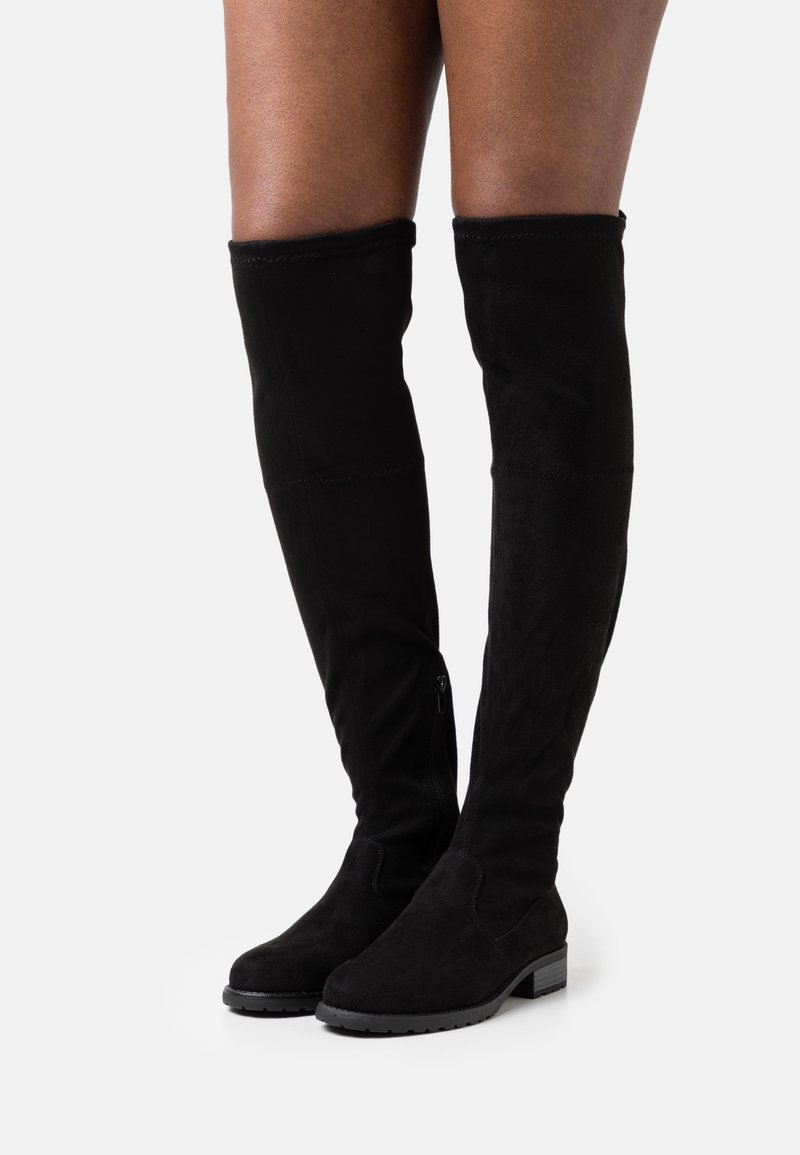Jambes portant des bottes plates noires au-dessus du genou avec des petits talons blocs et des fermetures éclair latérales, sur un fond clair uni.