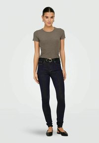 Jovem mulher de pé, vestindo uma blusa corta de malha castanha, jeans skinny escuros com cinto preto e sapatilhas pretas, contra um fundo branco.