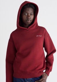 Rote Kapuzen-Sweatshirt aus weichem Stoff, mit einer Kängurutasche, gerippten Bündchen und Saum sowie einem kleinen weißen Logo auf der Brust.
