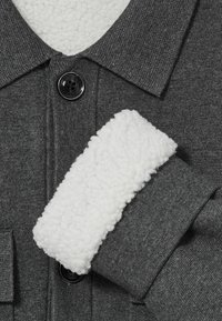 Cămașă din țesătură gri cu un buton negru și guler și manșete din fleece alb, având o suprafață texturată și moale.