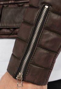 Manica di giubbotto in pelle marrone con un design texturizzato e trapuntato e una zip argentata; presenta un rivestimento in tessuto nero attorno alla zip.