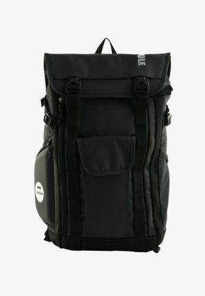 Sac à dos noir en tissu résistant avec un design à rabat, plusieurs poches, des sangles réglables et un patch circulaire.