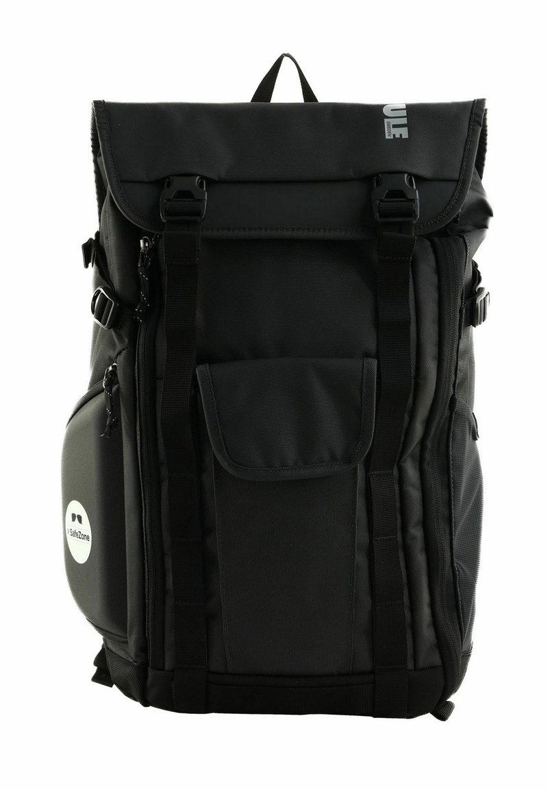 Sac à dos noir en tissu résistant avec un design à rabat, plusieurs poches, des sangles réglables et un patch circulaire.