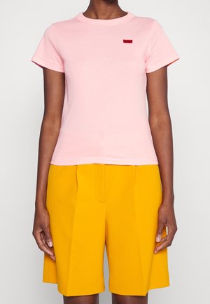 Person trägt ein schlichtes, hellrosa kurzärmeliges T-Shirt und hoch taillierte, leuchtend gelbe, maßgeschneiderte Shorts und steht vor einem schlichten Hintergrund.