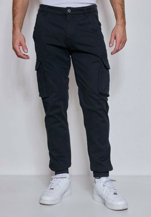 Homme portant un pantalon cargo noir avec des poches et des baskets blanches, debout contre un fond clair uni.