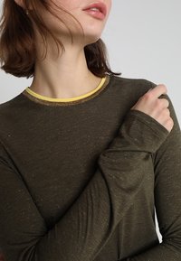 Olijfgroene top met lange mouwen, een ronde gele kraag en subtiele glitters in de stof, met aansluitende mouwen en een zachte textuur.