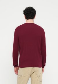 Μακρυμάνικο πουκάμισο σε βυσσινί με ριμπ ύφανση, στρογγυλή λαιμόκοψη και εφαρμοστά μανσέτα, συνδυασμένο με ανοιχτό μπεζ παντελόνι cargo με πλαϊνές τσέπες.