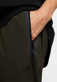 Olivengrønne Nike joggers med en lynlåslomme, sorte accenter og en glat tekstur. Inkluderer et diskret logo på siden.