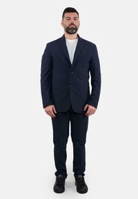 Completo da uomo blu con giacca su misura e pantaloni abbinati, indossato sopra una t-shirt bianca. Presenta rever standard e due bottoni. Sneakers nere.
