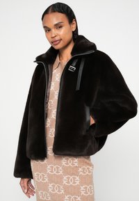 Casaco de pelúcia castanha com colarinho alto, fecho de correr à frente e detalhes em couro negro. O design inclui bolsos laterais e uma textura macia.