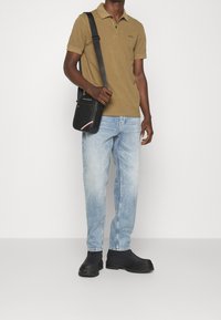 Brun polo-tröja med logotyp, ljusblå jeans och svart axelväska. Svarta stövlar med strukturerade sulor. Enkel och funktionell design.