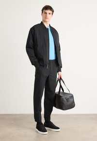 Zwarte bomberjack, lichtblauw T-shirt, zwarte tapered broek, zwarte sneakers met witte zolen, houdend een zwarte sporttas. Effen achtergrond.