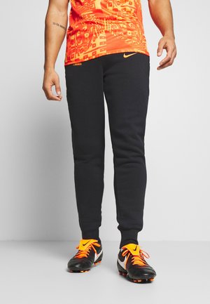 Osoba w czarnych spodniach dresowych Nike, pomarańczowej sportowej koszulce z wzorem oraz czarnych korkach Nike z pomarańczowymi sznurowadłami i dodatkami, stojąca na szarym tle.