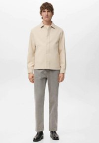 Giacca camicia beige con colletto, indossata sopra una maglietta chiara, abbinata a jeans grigi e stivaletti neri. Texture liscia, vestibilità comoda.