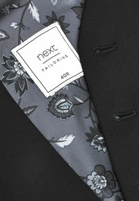 Schwarze maßgeschneiderte Jacke mit einem Innenfutter in grauem Blumenmuster. Das Etikett trägt die Aufschrift "next TAILORING" mit der Größe "40R".
