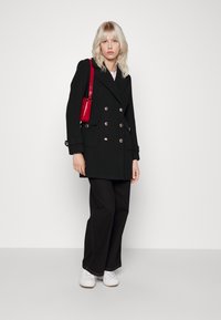 Manteau noir croisé avec des boutons dorés, associé à un pantalon noir à jambes larges et des baskets blanches. Accessoirisé avec un sac à main rouge.