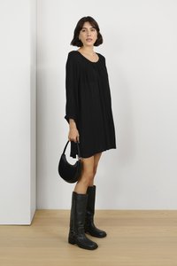 Schwarzes, langärmliges Kleid mit gefaltetem Design. Kombiniert mit knielangen schwarzen Lederstiefeln und einer kleinen, geschwungenen schwarzen Handtasche.