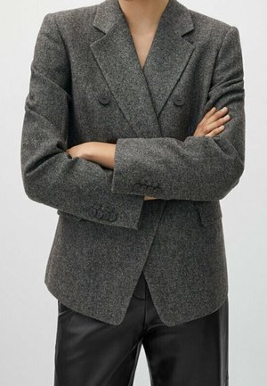 Persona con le braccia incrociate che indossa una giacca grigia doppiopetto e pantaloni di pelle neri su uno sfondo neutro.