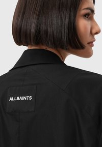 Giacca nera con un'etichetta logo visibile con la scritta "ALLSAINTS" sul retro. Il materiale sembra liscio, con linee di design affilate e sartoriali.