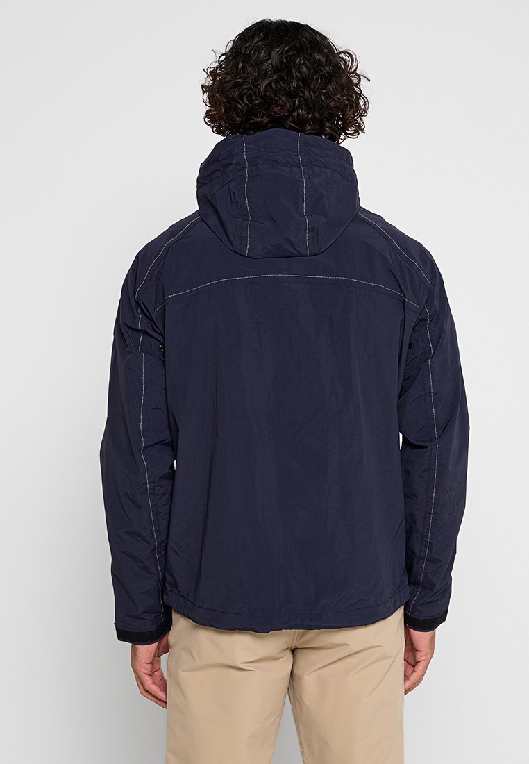 Uomo con capelli ricci e scuri, che indossa una giacca a vento blu navy con cappuccio e pantaloni beige, in piedi di spalle su uno sfondo bianco puro.