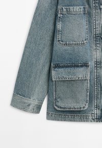 Spijkerjack in lichtblauw, met twee grote voorkruippockets, rechte snit en zichtbare stiksels langs de randen.