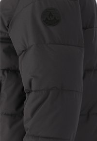 Sort pufferjakke med polstrede sektioner og en rund sort logo-patch på skulderen. Stoffet ser glat og vandafvisende ud.