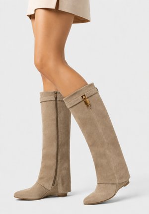 Beine, die beige Wildleder-Kniestiefel mit seitlichen Reißverschlüssen und goldenem Schlossdetail tragen, kombiniert mit einem beigen Rock vor weißem Hintergrund.