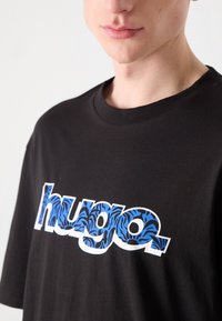 Musta puuvillainen t-paita, jossa on valkoisella ääriviivalla oleva "hugo"-logo, jonka sisällä sinimusta tiikerinraidoituskuvio. Lyhyet hihat.