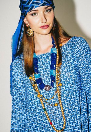 Junge Frau mit einem blauen, gemusterten Oberteil und Kopftuch, geschmückt mit mehreren bunten Perlenketten und großen dekorativen Ohrringen.