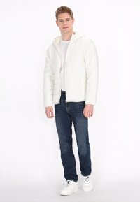 Witte gewatteerde jas met een capuchon, ritsluiting, twee zijzakken, gecombineerd met donkerblauwe jeans en witte sneakers, staand tegen een effen achtergrond.