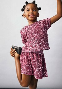 Set strălucitor format din top și fustă roz cu paiete, cu mâneci scurte și croială lejeră. Pantofi negri și o pose jucăușă îi accentuează aspectul vibrant.