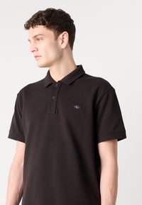 Svart poloskjorte med teksturert vev, med en tre-knappers placket og et lite merketag på brystet. Korte ermer.