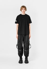 Camiseta de algodón negra con lettering texturizado, combinada con pantalones cargo negros con cremalleras y correas, finalizando con calzado negro.